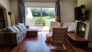 Ein modernes Wohnzimmer mit einer Couch, einem Holzstuhl, einem Fernseher und einer Schiebetür zu einer Terrasse mit Gartenmöbeln.