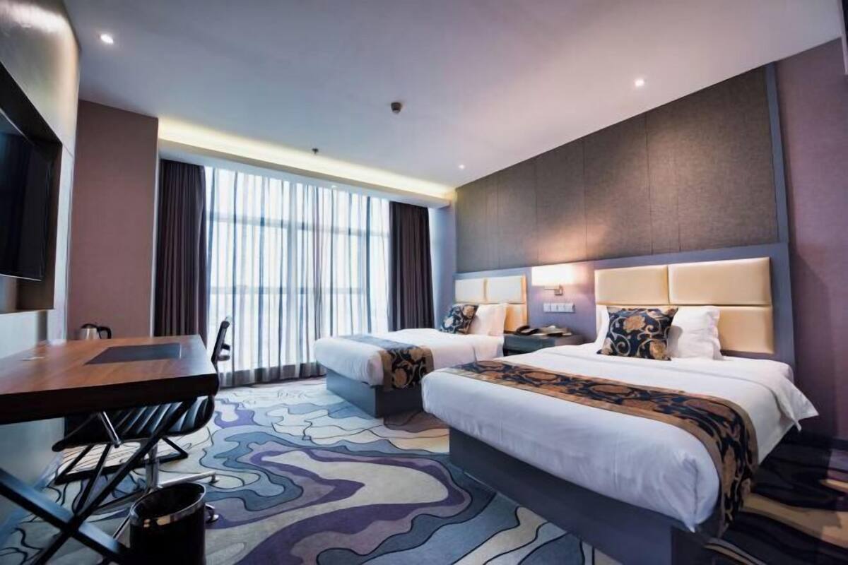 A hotel room with two beds, a desk, and a large window with curtains.