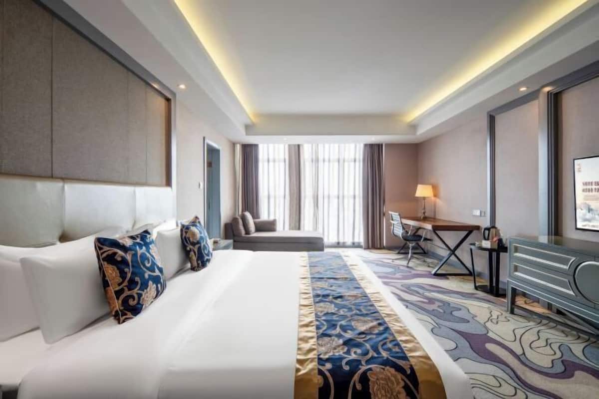 A hotel room with a large bed, a desk, and a chair. There is a window with curtains and a view of the outside.