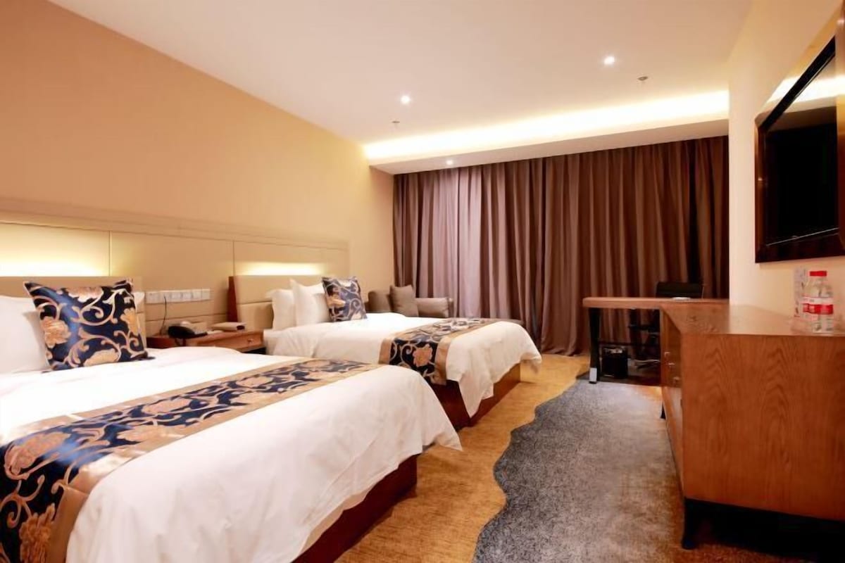 A hotel room with two beds, a desk, a television, and a large window with curtains.