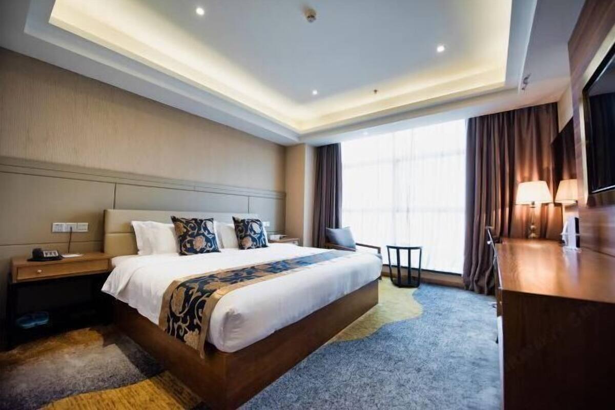 A modern hotel room with a large bed, a nightstand, a desk, and a view of the outdoors.