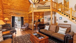A log cabin interior with a wooden staircase, a living room with a sofa and armchairs, a dining area, and a kitchen.