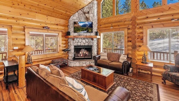 A log cabin interior with a stone fireplace, wooden furniture, and large windows.