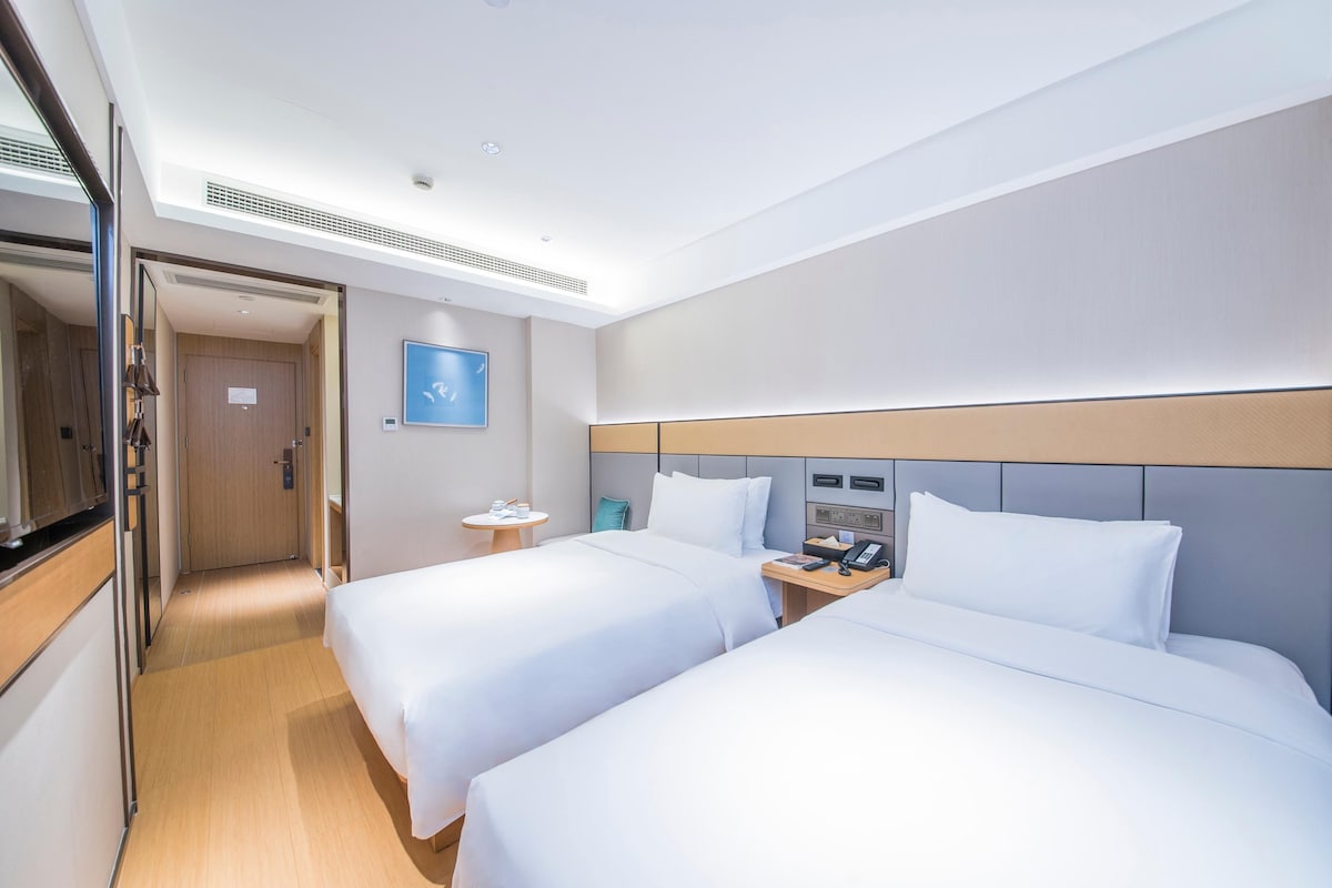 A hotel room with two beds, a bedside table with a phone, a small round table with a lamp, and a door leading to another room.