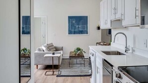 A modern kitchen with white cabinets, a stainless steel sink, and a built-in oven. There is a gray sofa and a small table with a plant in the living area. - Sosuite | 1BR Apt w W/D, Gym, Lounge, Park View (Philadelphia)