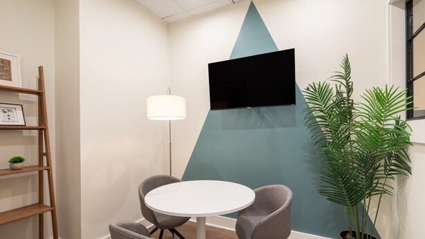 A conference room with a white table, two grey chairs, a floor lamp, a TV mounted on a teal wall, a bookshelf, and a potted plant. - Sosuite | 1BR Corner Loft w Park View, Gym, Lounge (Philadelphia)