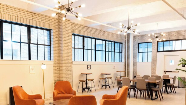 A spacious room with large windows, a brick wall, and modern furniture including orange chairs and tables. - Sosuite | 2BR Apt w W/D, Gym, Lounge (Philadelphia)