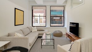 A modern living room with a sofa, coffee table, and TV. There are two windows with views of buildings outside. - Sosuite | 2BR Apt w W/D, Gym, Lounge (Philadelphia)