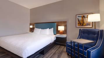 A hotel room with a bed, a nightstand with a lamp, a blue armchair, and a framed picture on the wall.