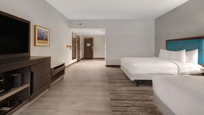Habitación de hotel con un televisor de pantalla plana grande, una cama con ropa blanca, un cabecero de madera y una alfombra a rayas.