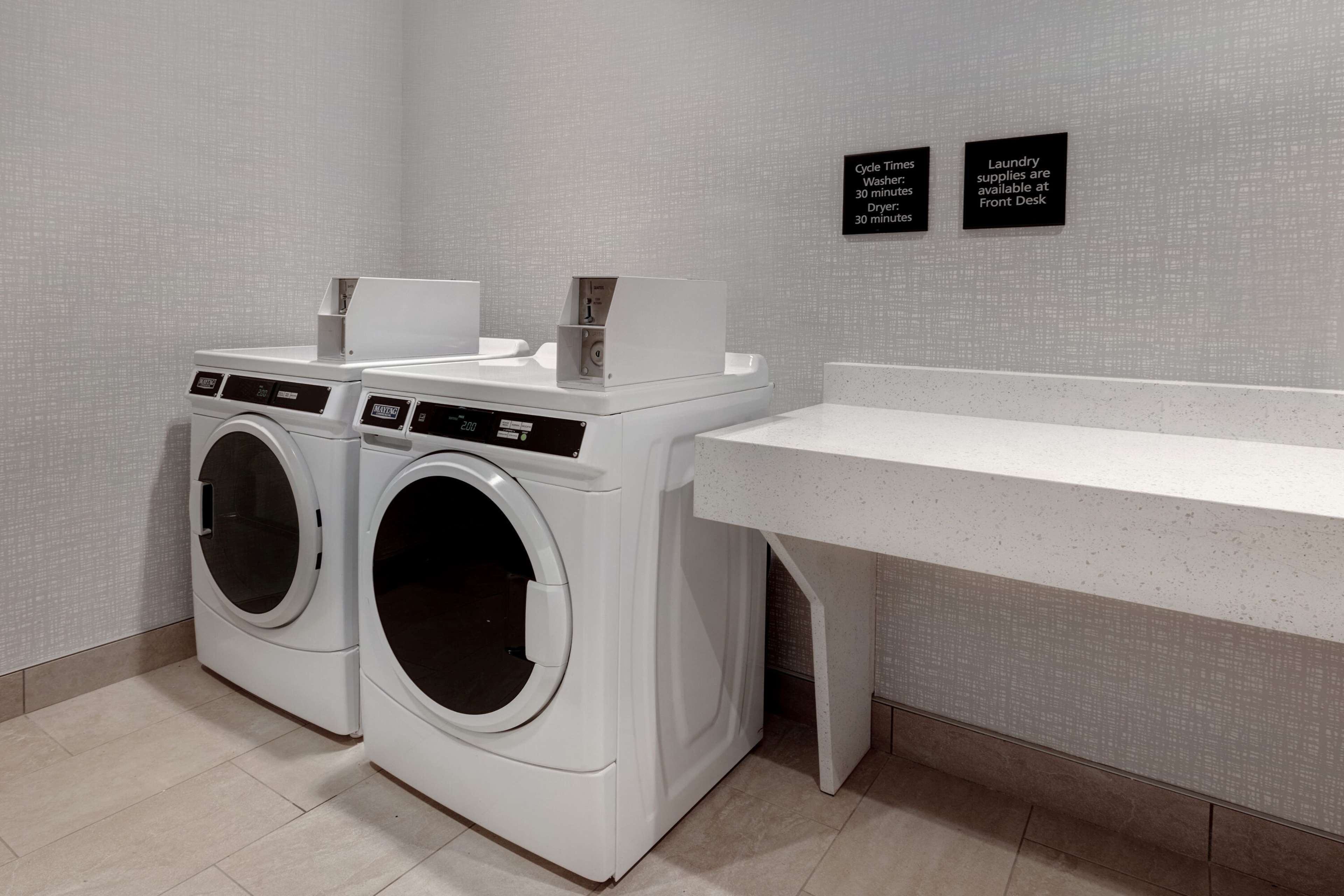 A laundry area with two front-loading washing machines, a drying station, and signs indicating cycle times and availability of laundry supplies.
