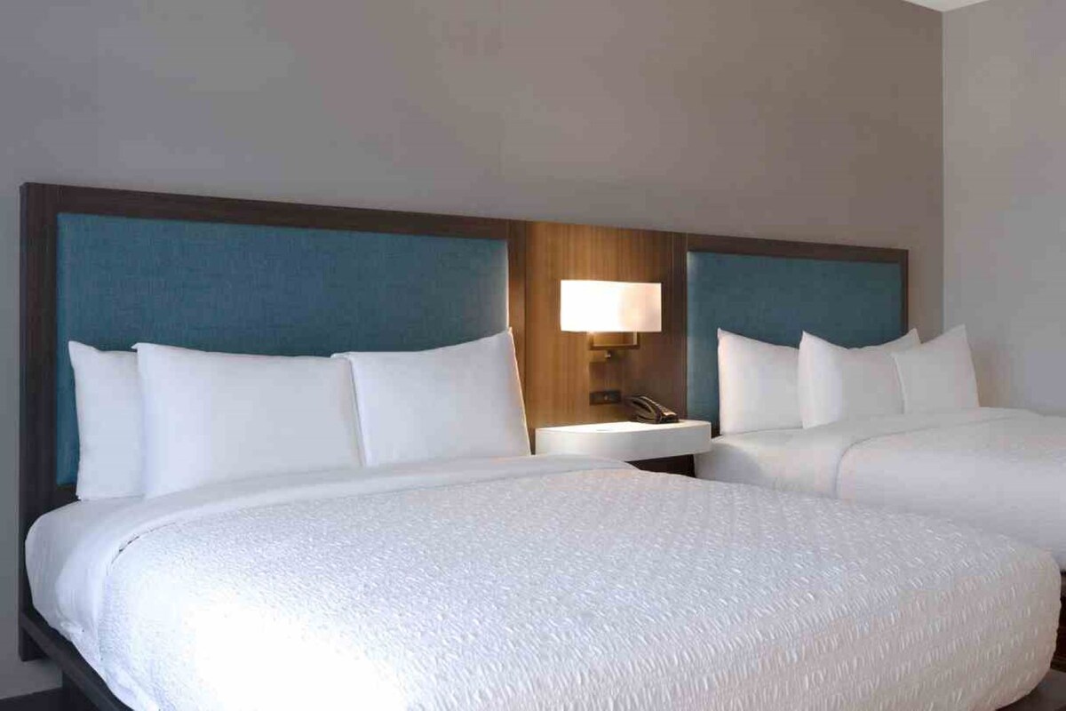 A hotel room with two beds, a nightstand with a lamp, and a small table.