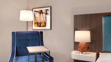 A hotel room with a blue upholstered chair, a small table, a wall-mounted lamp, and a framed picture.