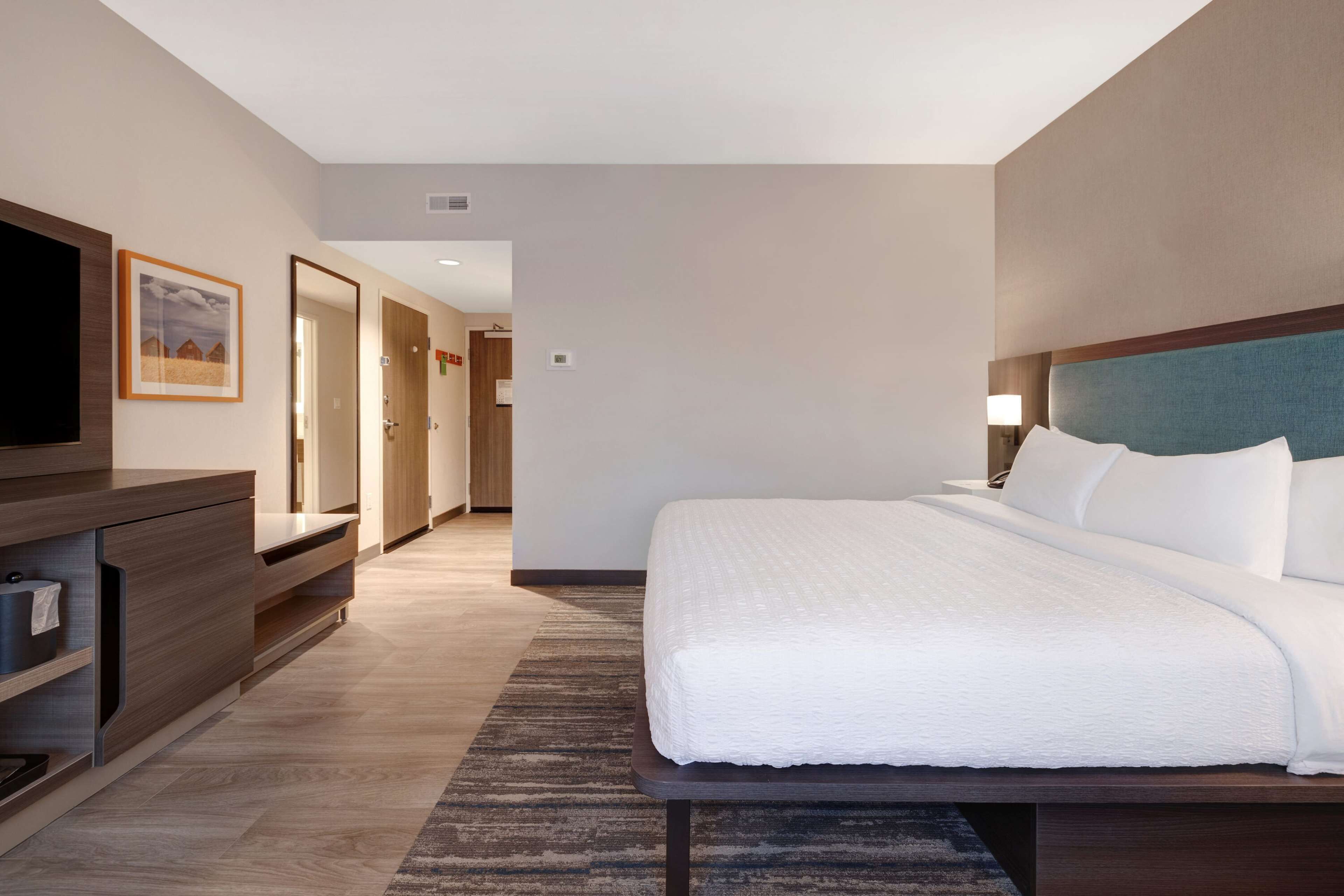 A hotel room with a large bed, a TV, a wooden dresser, and a hallway leading to another room.