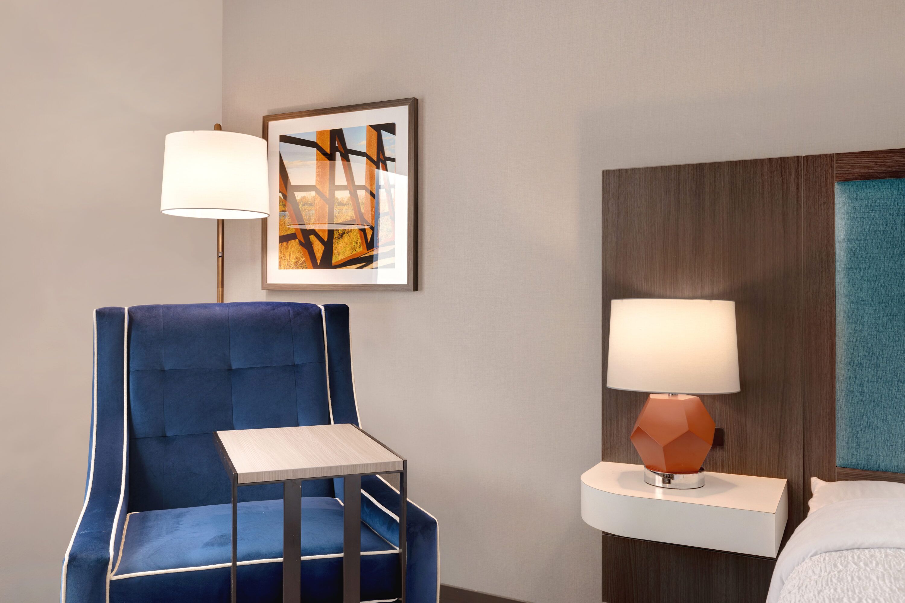 A hotel room with a blue upholstered chair, a small wooden table, a white bedside table with a lamp, and a framed abstract painting on the wall.