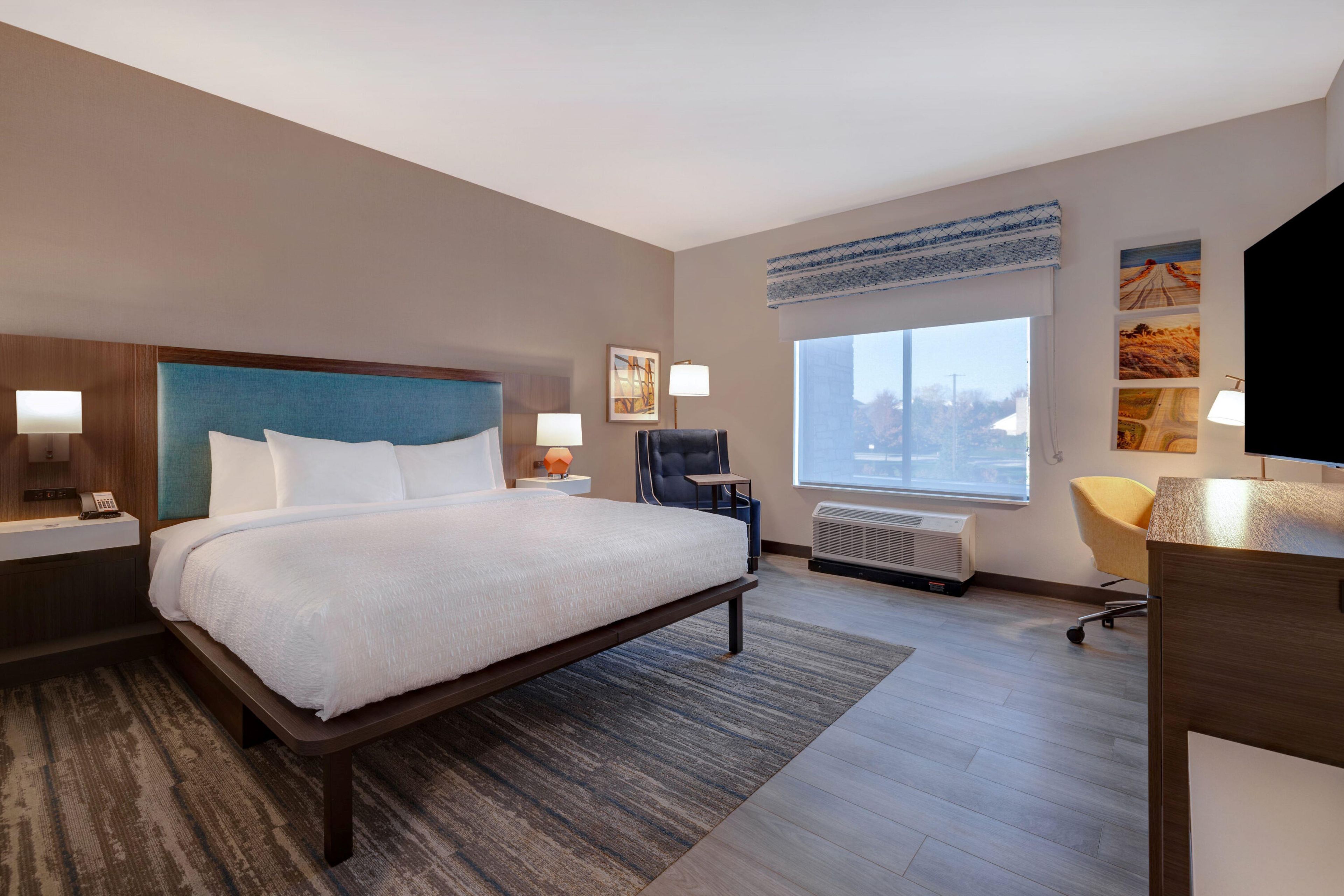 A hotel room with a large bed, a desk, a chair, a window with a view, and artwork on the walls.