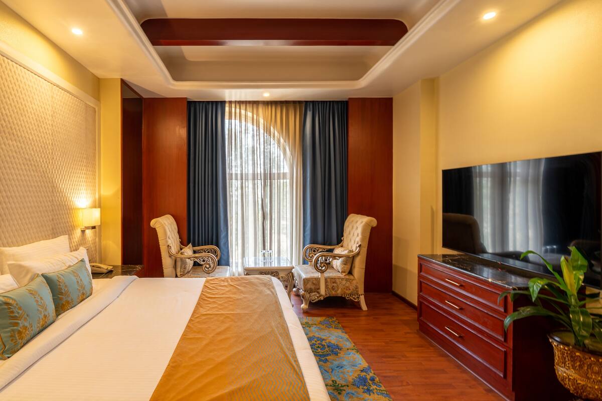 A hotel room with a large bed, two armchairs, a TV, a dresser, and a large window with curtains.