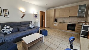 A modern living room with a grey sofa, a white coffee table, and a blue ottoman. A kitchen with wooden cabinets and a tiled backsplash is visible in the background. - Regarda - "Costa del Sole" with swimming pool and terrace (Lazise)