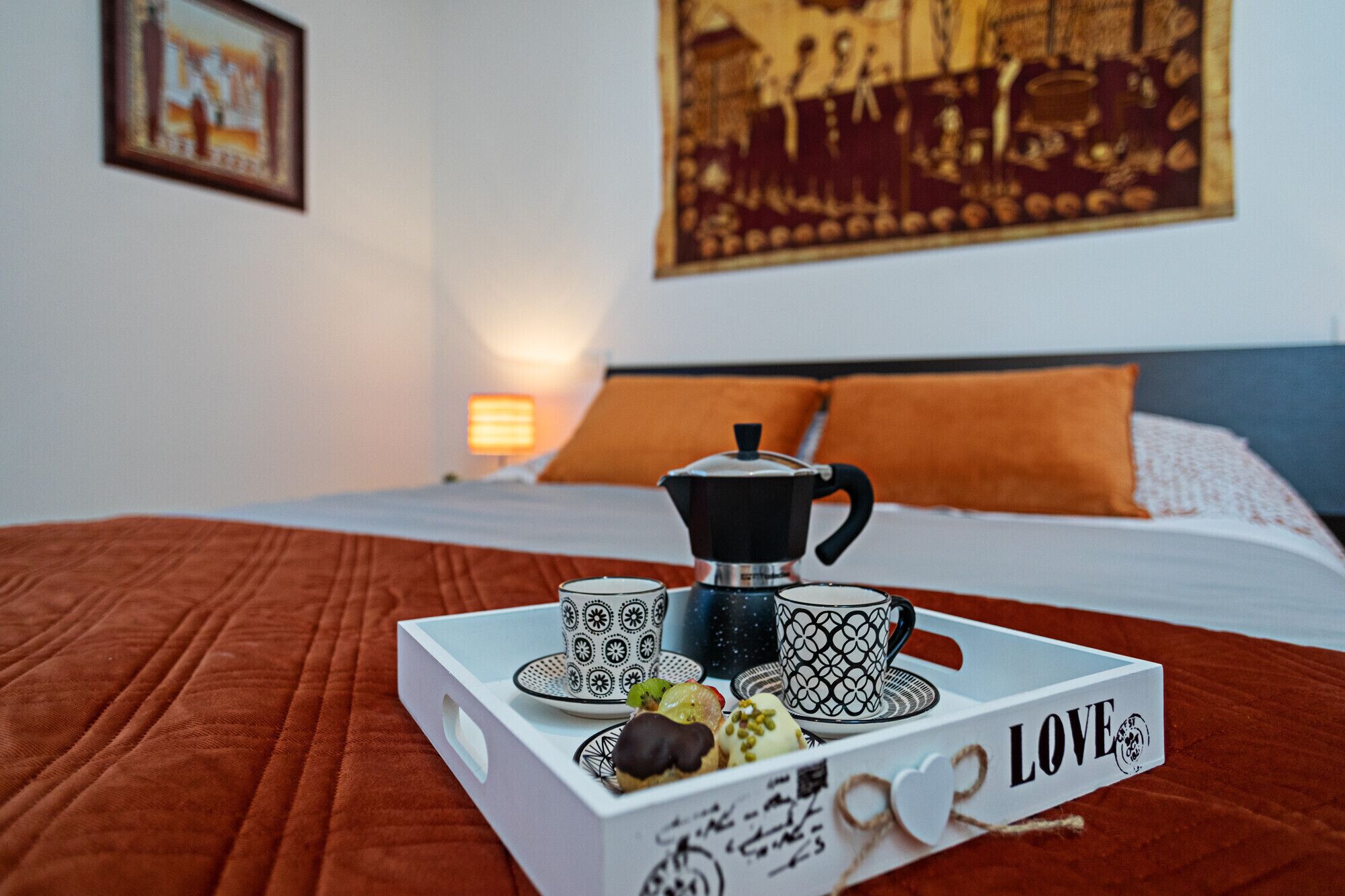A neatly made bed with a white comforter and an orange pillow, a coffee pot, and a tray with a coffee cup and pastries on a white tray with the word "LOVE" on it.