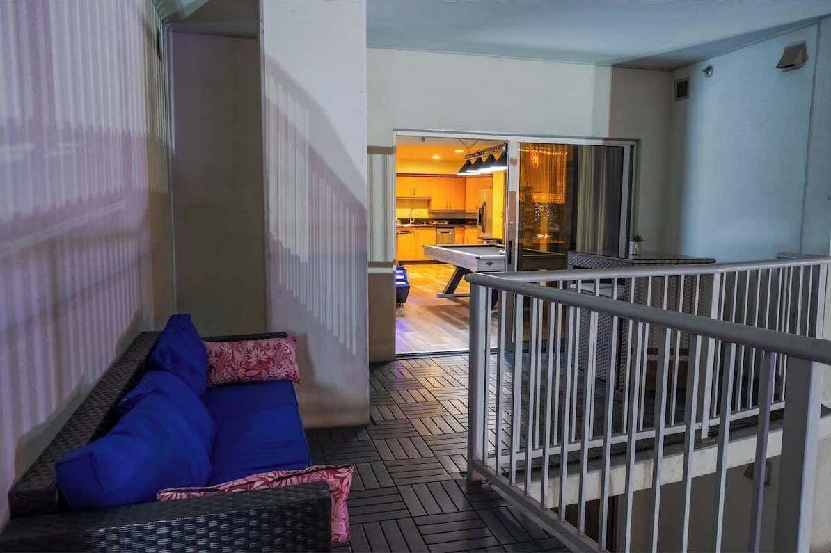 Un balcón con sofá azul y una pequeña mesa, barandilla de metal y vista hacia una cocina.