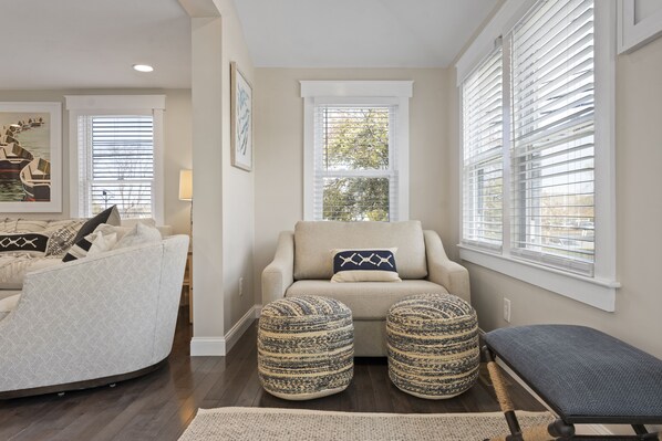 A cozy living room with a sofa, ottomans, and a bench. There are two windows with blinds, a painting on the wall, and a lamp.