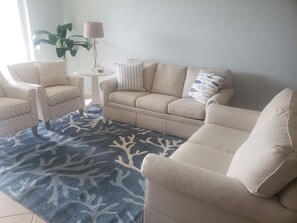 Relax in this inviting living space with a beige sofa, armchair, and coffee table, complemented by a colorful rug and a potted plant.