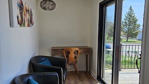 A modern living room with a wooden desk, two black leather chairs with blue cushions, a wicker pendant light, and a large sliding glass door leading to a deck.