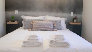 Una cama impecablemente arreglada con toallas blancas dobladas, una almohada gris estampada y dos mesitas de noche con plantas en macetas.