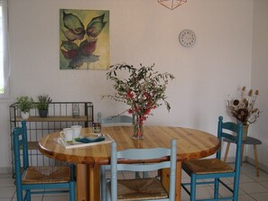 A dining area with a round wooden table, four blue chairs, a vase with flowers, a clock on the wall, and a painting above the table. - Charming air-conditioned gîte, Chauzon Ardèche méridionale (Chauzon)