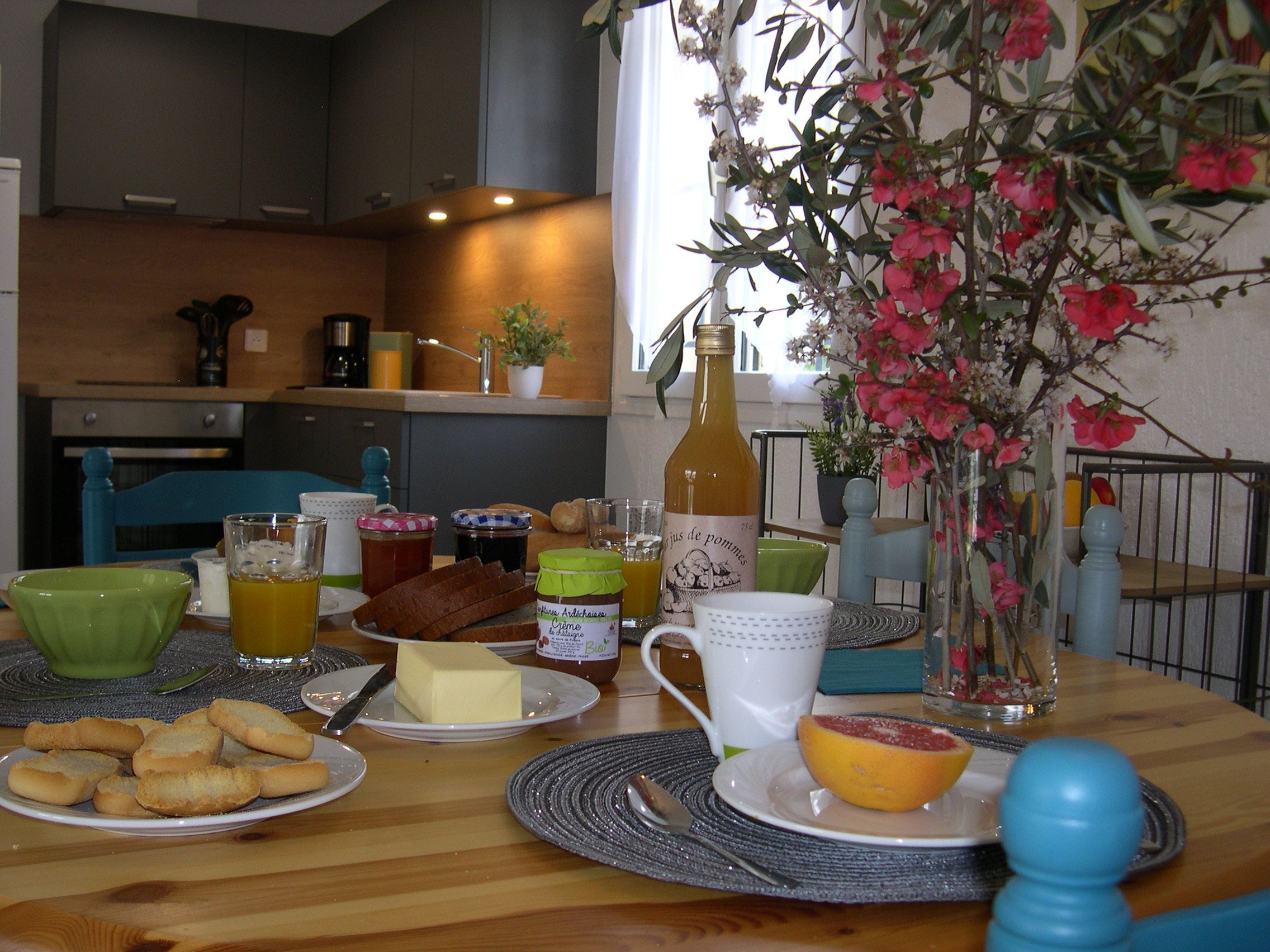 A breakfast table with various food items, a bottle of olive oil, and a vase with flowers.