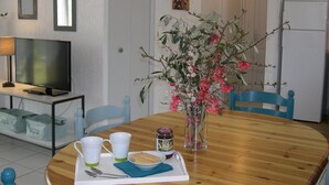 A dining area with a wooden table, two chairs, a vase with flowers, a tray with a plate of crackers, two cups, a jar of jam, and a television on a stand. - Charming air-conditioned gîte, Chauzon Ardèche méridionale (Chauzon)