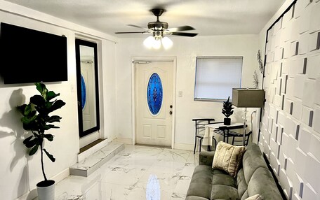 A living room with a sofa, a dining table, a ceiling fan, a TV mounted on the wall, and a door with a blue glass window.