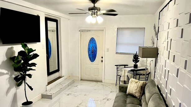 A living room with a sofa, a dining table, a ceiling fan, a TV mounted on the wall, and a door with a blue glass window. - Miami Gardens Comfort Suite by Hard Rock Stadium (Miami Gardens)