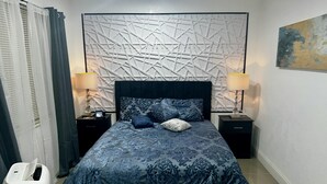 A bedroom with a bed, two nightstands, two lamps, a wall with a geometric pattern, and a trash bin. - Miami Gardens Comfort Suite by Hard Rock Stadium (Miami Gardens)