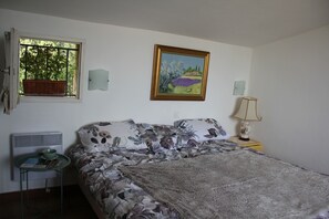 A bedroom with a bed, a window with a view of trees, a painting on the wall, and a bedside table with a lamp.