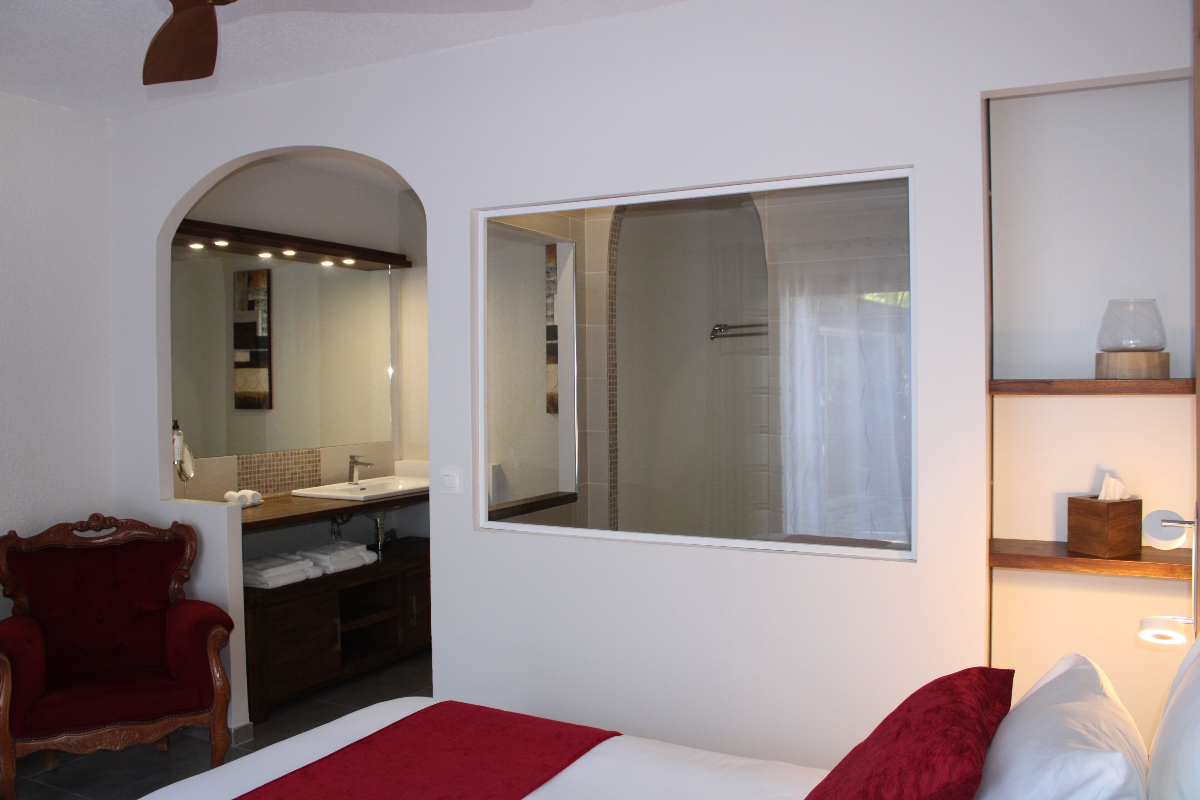 A hotel room with a large mirror, a vanity with a sink, a shower, a red armchair, and a bed with white bedding.