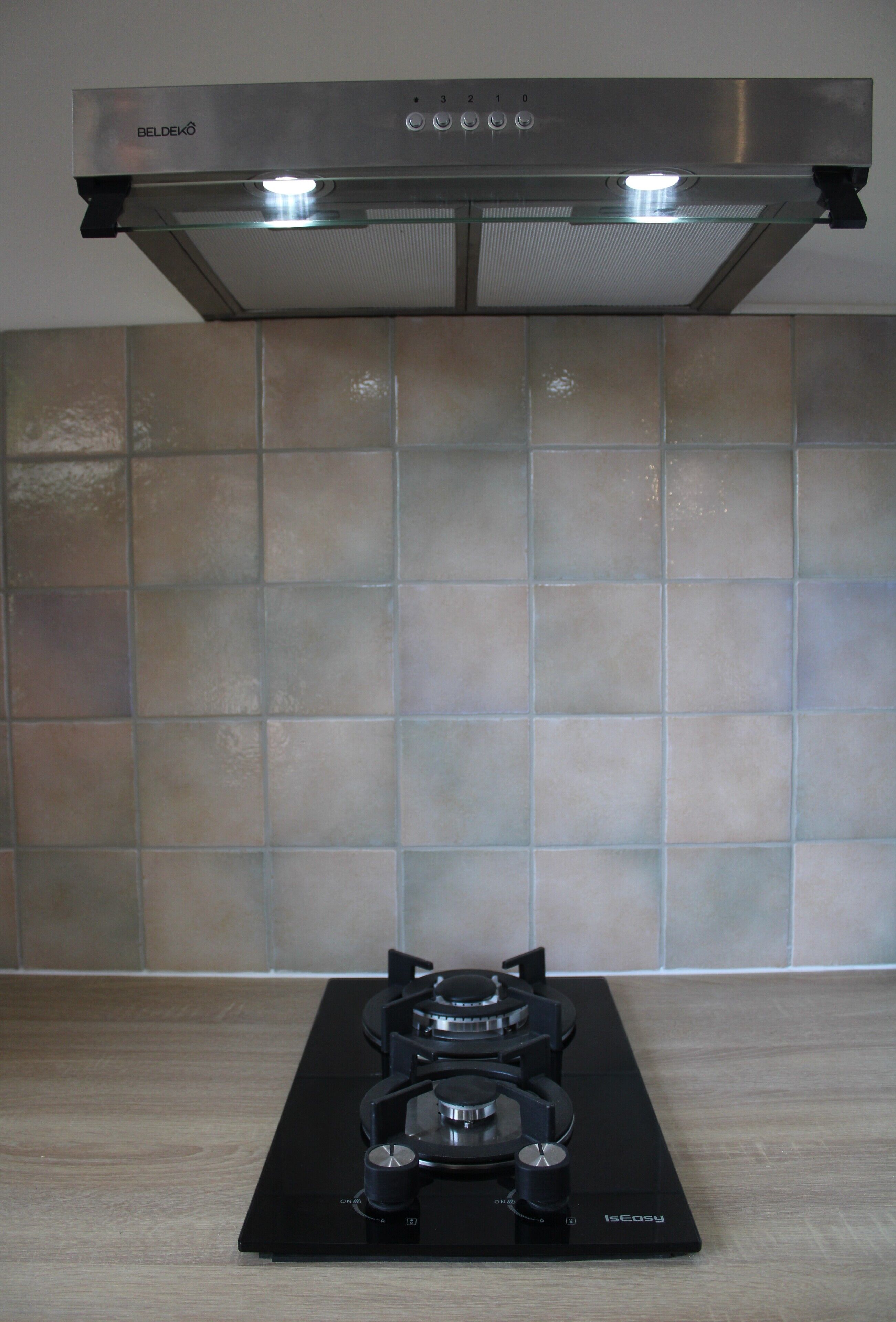 Een keuken met een roestvrijstalen afzuigkap, een zwart inductiekookplaat en een beige betegelde achterwand.