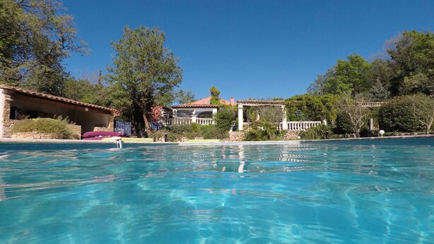Une piscine aux eaux cristallines, bordée de pierres, entourée d’une végétation luxuriante et d’un bâtiment doté d’un toit en tuiles.