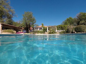A clear swimming pool with a stone border, surrounded by lush greenery and a building with a tiled roof. - Les Secrets du Defends (Forcalqueiret)