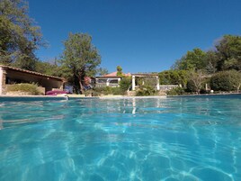 Piscina cristalina con borde de piedra, rodeada de exuberante vegetación y un edificio con tejado de baldosas.