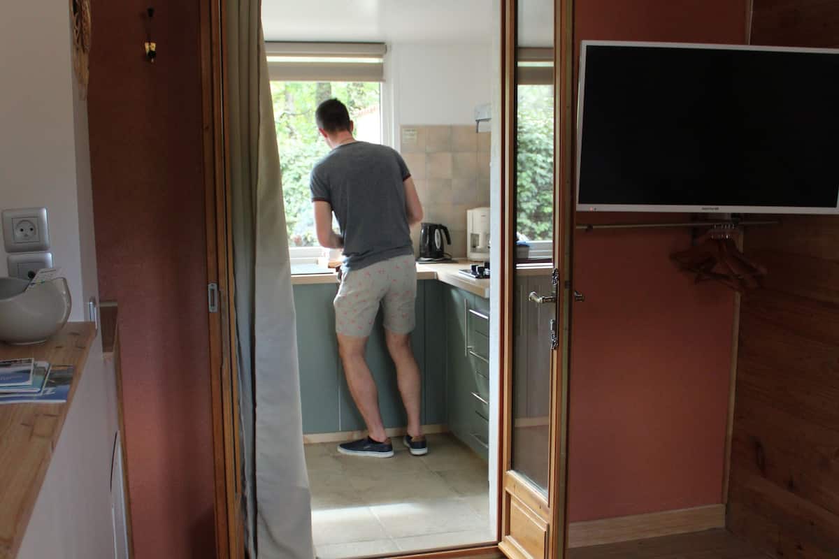 A bedroom with a bed, a wooden floor, a kitchen area visible through an open door, and a flat-screen TV mounted on the wall.