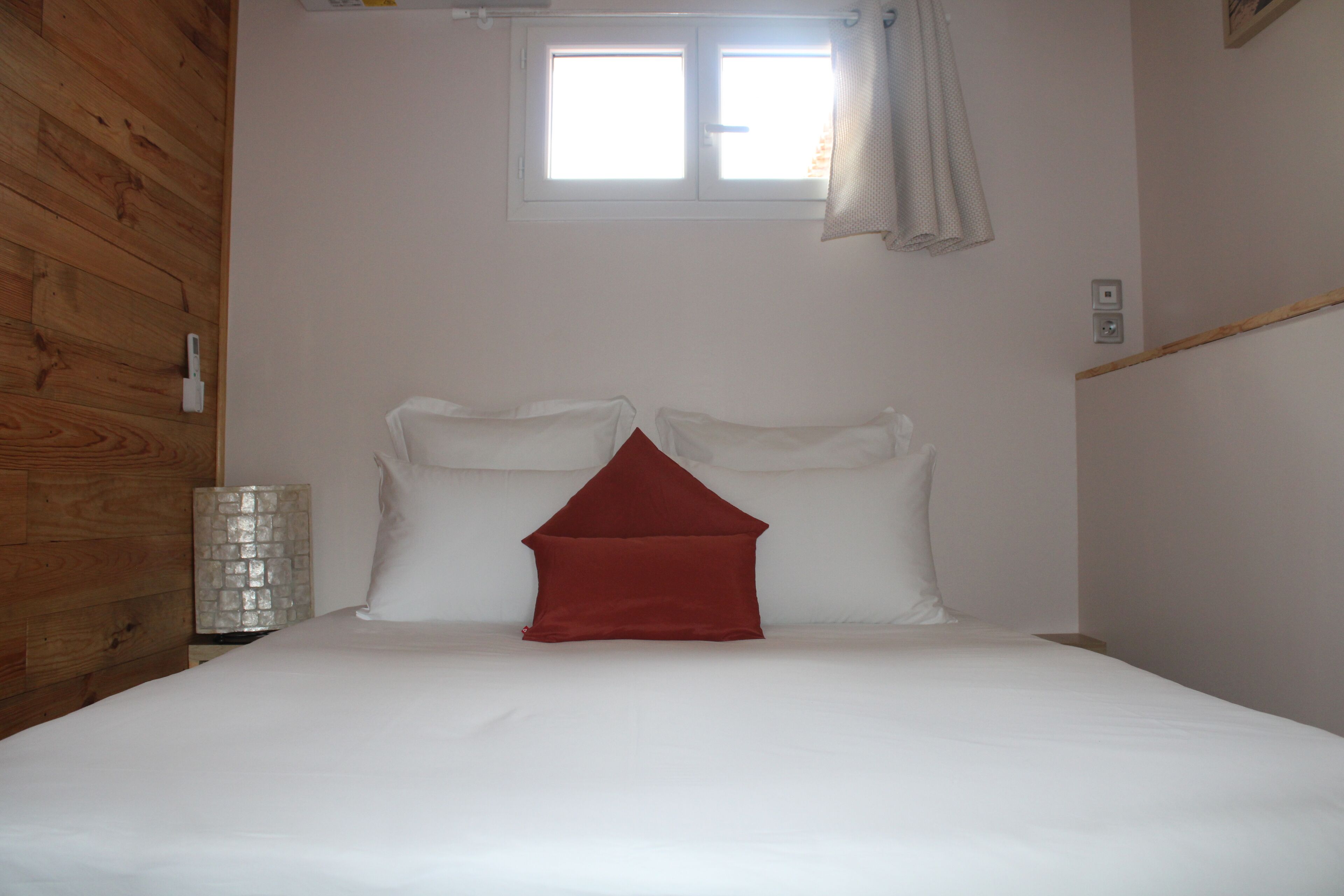 Una cama con ropa blanca y una almohada roja, una ventana con cortinas y una pared de madera.