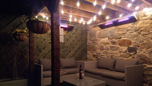 Un espace extérieur accueillant, avec un mur en pierre en arrière-plan, une jardinière suspendue et des guirlandes lumineuses.