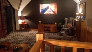 Un salon chaleureux avec une cheminée, un téléviseur fixé au mur, un fauteuil à carreaux et une table basse en bois.