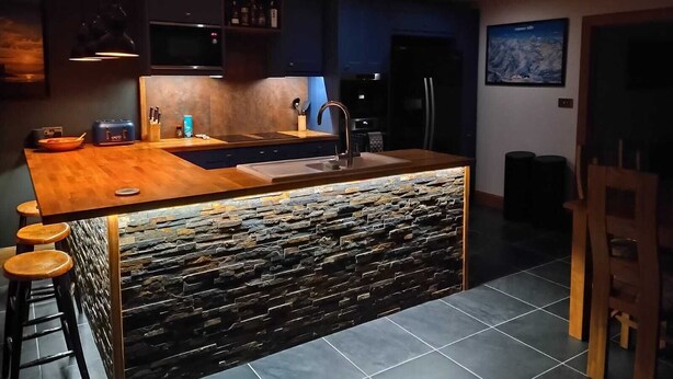 Une cuisine moderne dotée d’un îlot central, de meubles de couleur sombre et d’un mur d’accent en pierre.