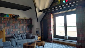 Un salon chaleureux avec un sol à carreaux, un canapé, deux petites tables basses en bois, une bibliothèque et une grande fenêtre avec des rideaux.