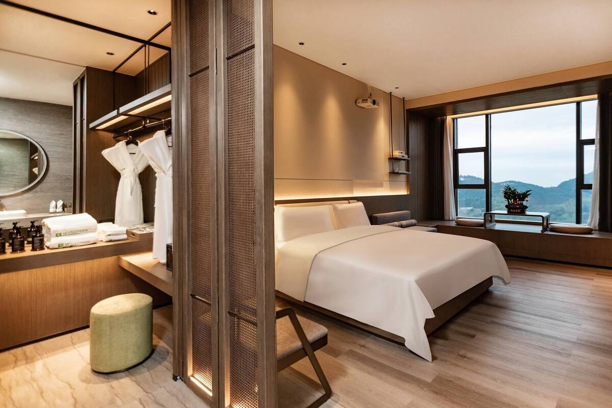 Une chambre d’hôtel moderne dotée d’un grand lit, d’une coiffeuse avec des articles de toilette, d’un dressing et offrant une vue sur les montagnes par la fenêtre.
