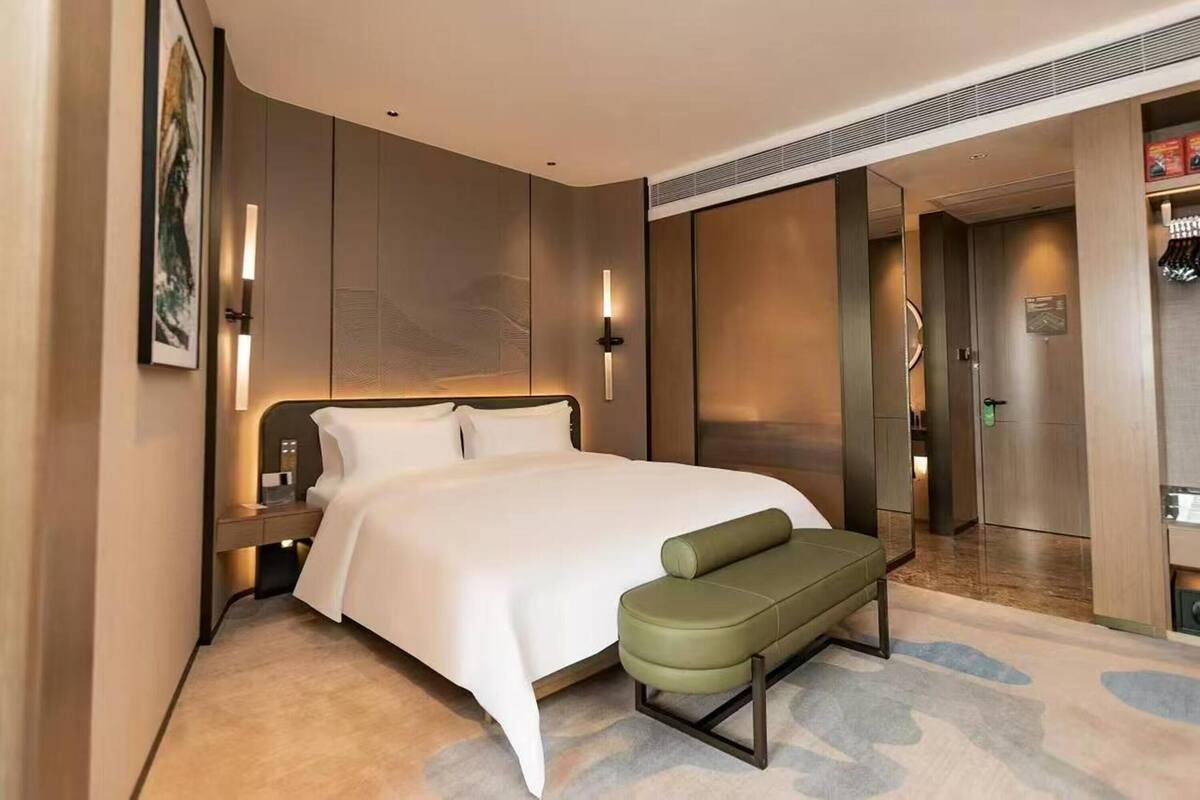 Une chambre d’hôtel moderne avec un grand lit, une banquette, une table de chevet et un luminaire fixé au mur.