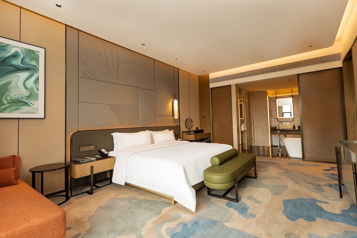 Une chambre d’hôtel moderne avec un grand lit, un bureau avec un téléphone, un banc, une salle de bain avec un miroir et un tableau au mur.