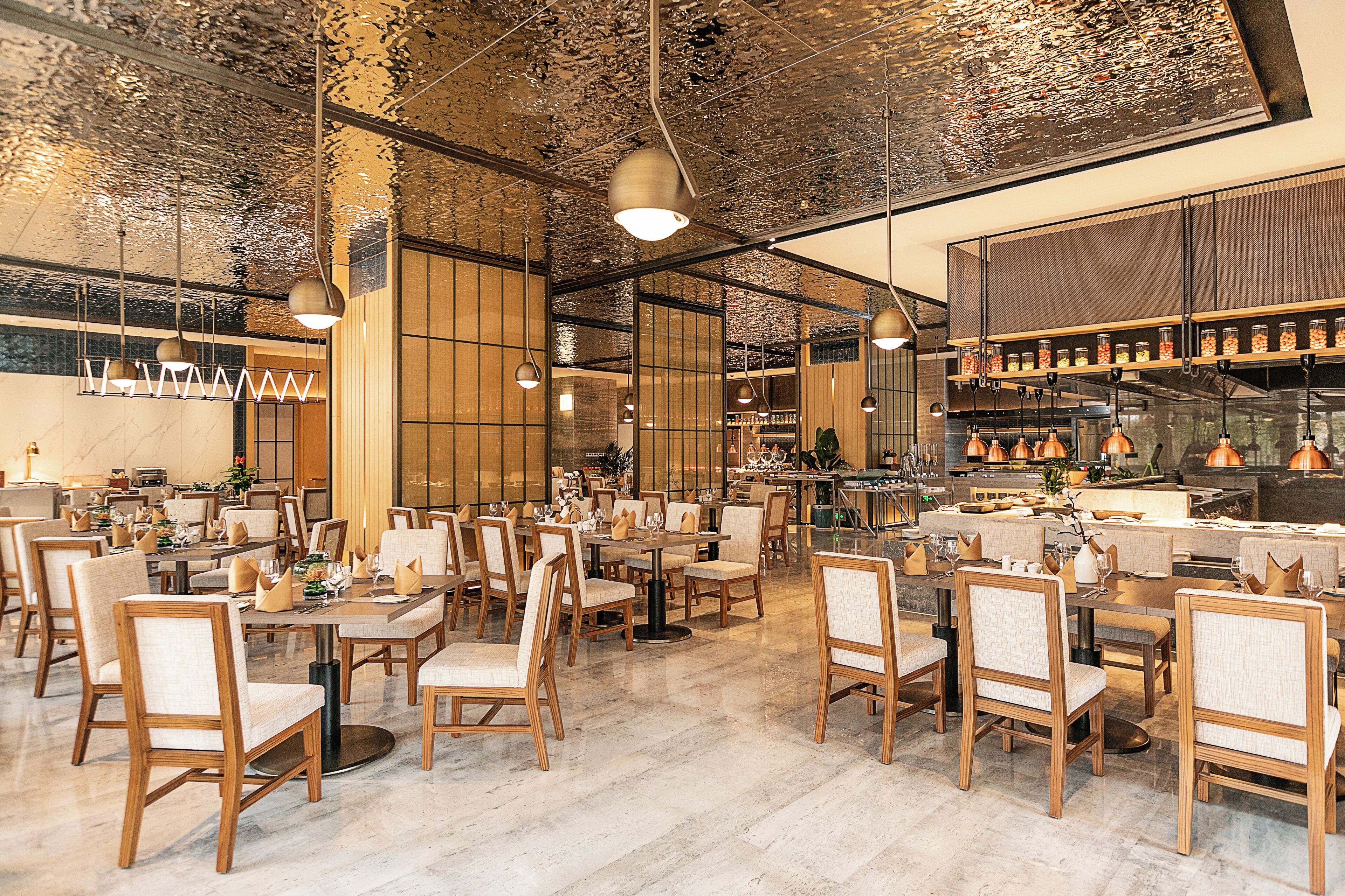 A spacious restaurant with wooden chairs and tables, a high ceiling with hanging lights, and a kitchen area visible in the background.